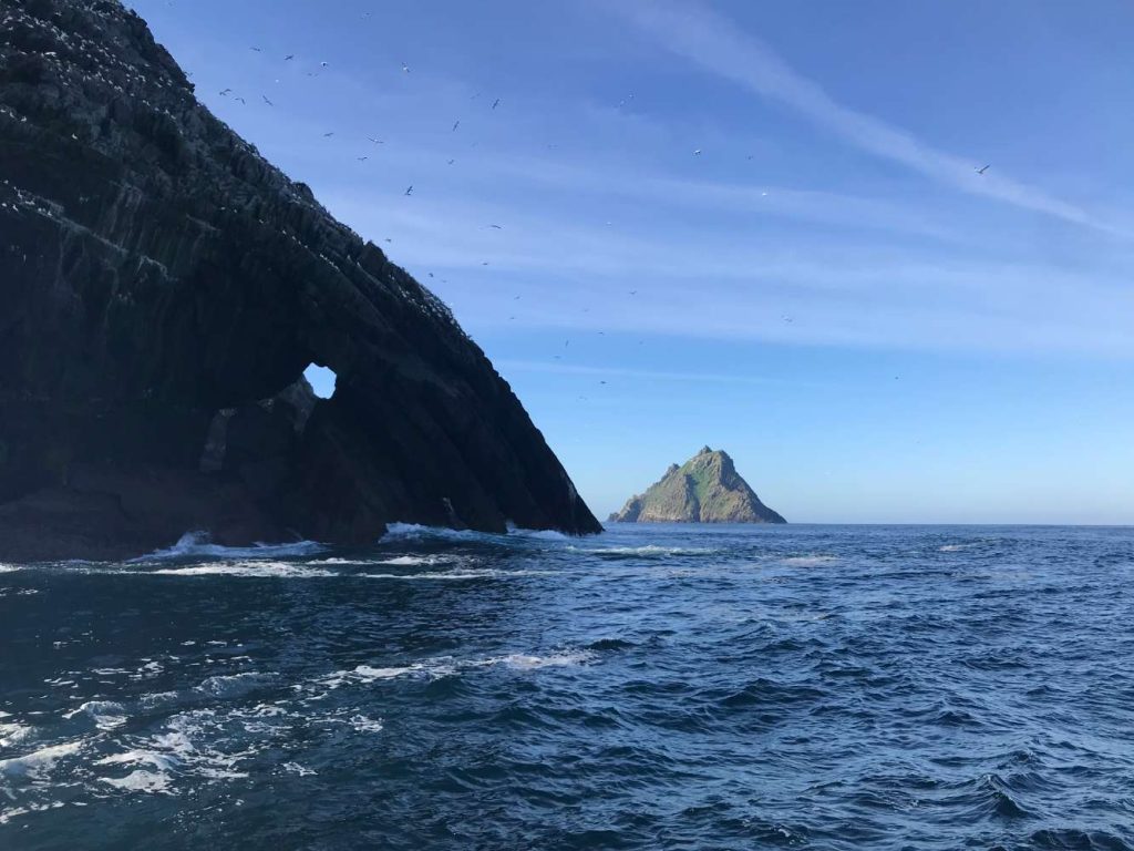 Little Skellig and the rugged coastline, viewed during Skellig Michael tours. Seabirds fly overhead. Waves crash against the rocks.